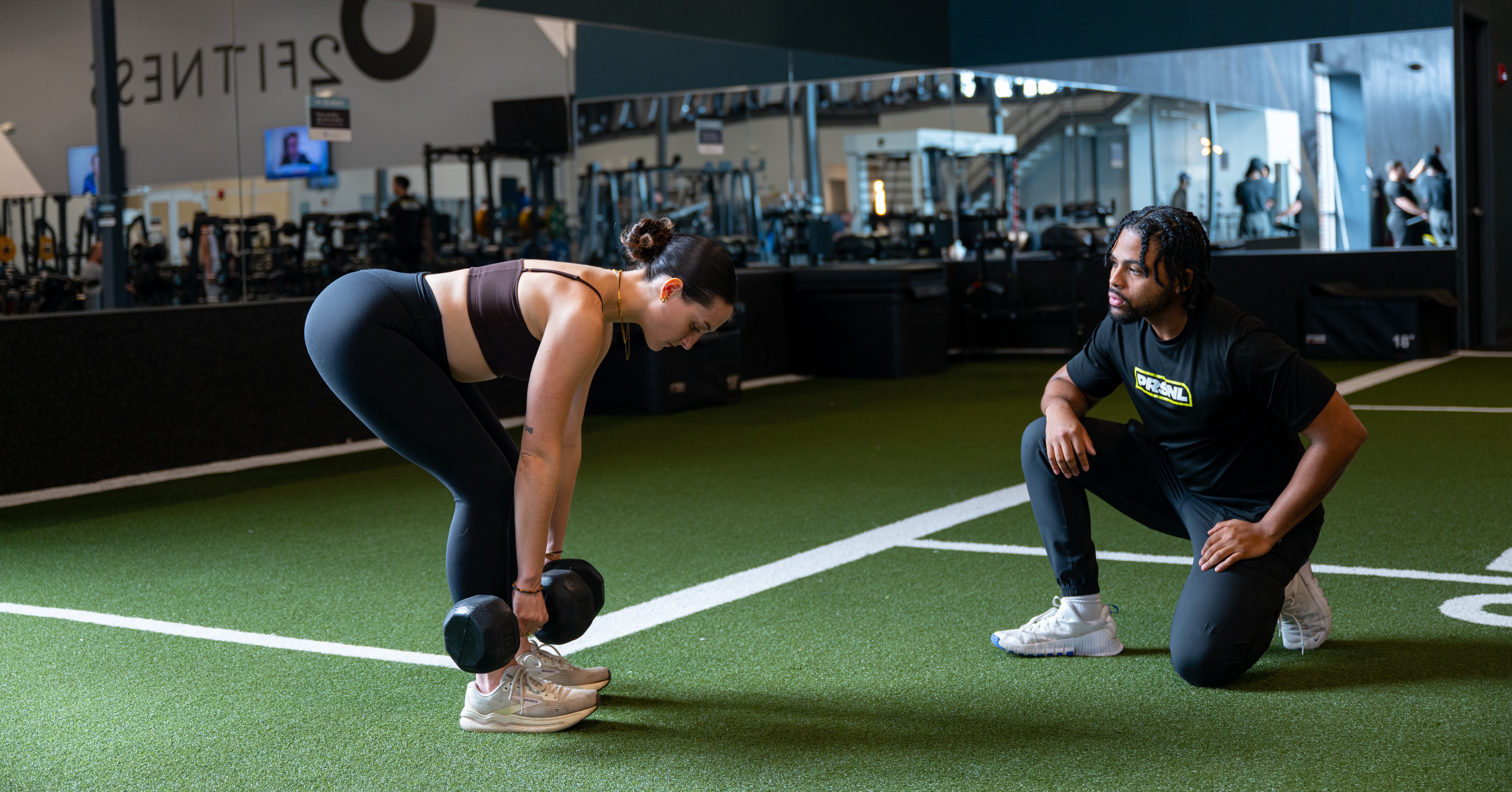 a woman performing dumbbell Romanian deadlifts with a PRSNL coach watching her form on the turf