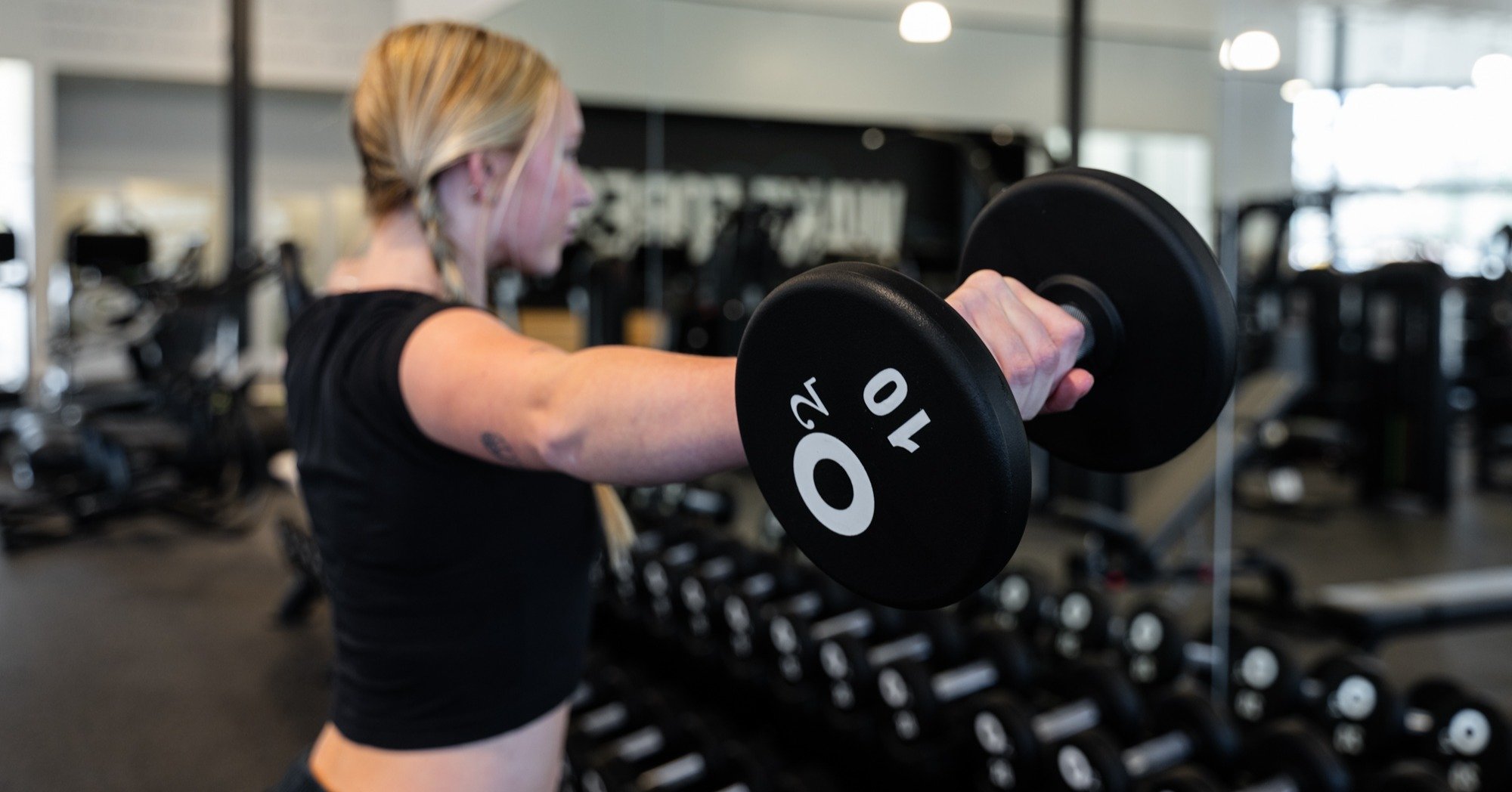 Young, blonde woman performing lateral raises in front of a mirror with a 10 pound dumbbell with an o2 fitness clubs logo 