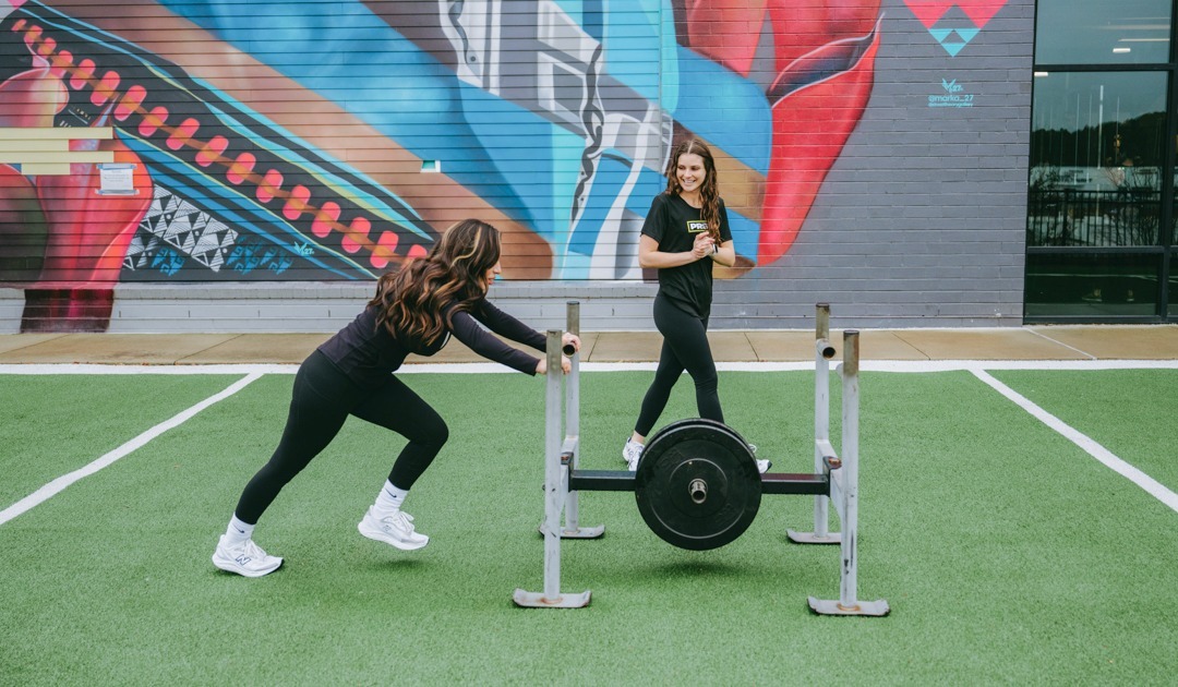 A woman performing a sled push on an outdoor turf while being encouraged by her personal trainer at O2 Fitness Clubs