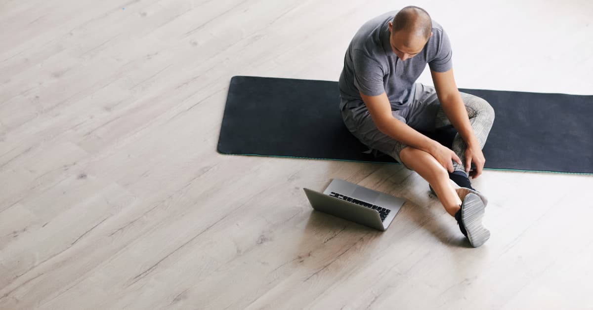 man stretching on a black yoga mat in front of his laptop in the gym